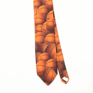 May include: A necktie featuring a repeating pattern of orange basketballs with black seams. The tie has a pointed tip and a tapered shape, displaying multiple basketballs of varying sizes across the fabric surface in a dense, overlapping arrangement.