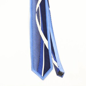Peut inclure: Une cravate présentant un motif géométrique de rayures verticales dans des tons bleu clair, bleu moyen et bleu marine foncé. Le design inclut une section à pois blancs et une rayure blanche incurvée. Une étiquette noire indique Made in Korea.