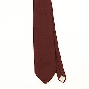 May include: A deep burgundy necktie featuring a subtle, repeating diagonal woven texture across the entire surface. The tie has a pointed bottom end and a visible white fabric lining at the narrow tip, showcasing the construction of the garment.