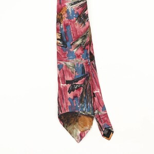 Op de afbeelding: Een stropdas met een abstract patroon in tinten roze, blauw, zwart en bruin. Het ontwerp bestaat uit penseelstreekachtige markeringen en geometrische vormen over de stof. De das loopt uit in een punt en bevat een klein zwart label.