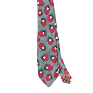 Peut inclure: Une cravate grise avec un motif répétitif de formes arrondies rouges, de carrés vert foncé et de petits carrés couleur crème. La cravate présente une extrémité pointue et une étiquette visible au dos près du bord inférieur du tissu.