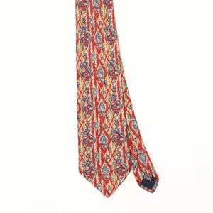 Op de afbeelding: Een stropdas met een felrode achtergrond en een herhalend patroon van goudkleurige diagonale lijnen die een ruitvormig raster vormen. Binnen elke ruit bevindt zich een gedetailleerd blauw en goud bloemmotief. De das heeft een puntig uiteinde.