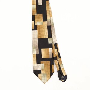 Peut inclure: Une cravate avec un fond noir et un motif géométrique composé de rectangles superposés dans des tons or, beige et crème. Chaque rectangle présente une texture fine en forme de grille sombre. La cravate a une extrémité pointue et une étiquette.
