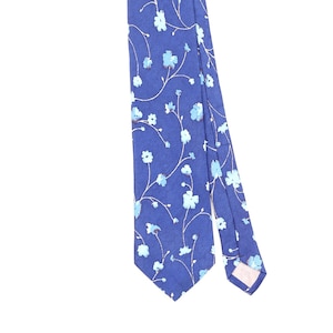 Pode incluir: Uma gravata azul com um padrão repetitivo de flores azul-claras ligadas por caules brancos finos e delicados. O tecido tem uma textura subtil em espinha e a gravata termina numa ponta com um forro de cor clara visível na parte inferior.