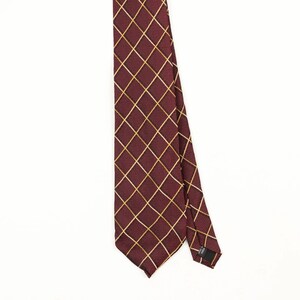 Könnte beinhalten: Eine Krawatte mit einer tiefburgunderroten Grundfarbe und einem sich wiederholenden goldenen Gittermuster aus diagonalen Linien. Der Stoff hat eine dezent strukturierte Oberfläche. Ein kleines schwarzes Etikett auf der Rückseite trägt den Text Made in Korea.