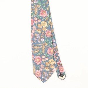 May include: A necktie featuring a dusty blue background with a repeating floral pattern of pink daisies, yellow pom-pom flowers, and golden wheat stalks. The tie includes a small black label on the back with white text indicating care instructions.