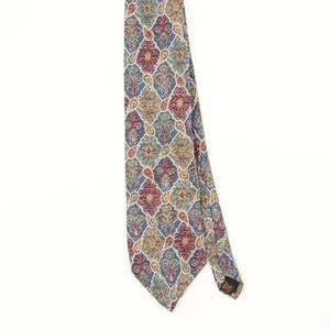 Può includere: Una cravatta con un motivo ripetitivo ispirato al damasco nei toni del blu, rosso, oro e crema. Il design è costituito da motivi interconnessi a forma di diamante con intricati dettagli floreali e paisley. Una piccola etichetta nera è attaccata sulla punta.