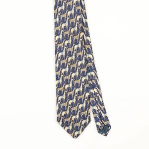 May include: A navy blue necktie featuring a repeating pattern of cream-coloured llamas or alpacas. Each animal is depicted in a standing position and wears a small, light blue saddle or blanket on its back. The fabric has a smooth, matte finish.