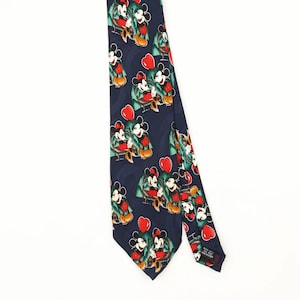 Pode incluir: Uma gravata azul-escura com um padrão repetitivo de personagens do Mickey Mouse sentados em bancos verdes, intercalados com formas de corações vermelho-brilhantes. A gravata tem uma extremidade pontiaguda e uma etiqueta preta visível.