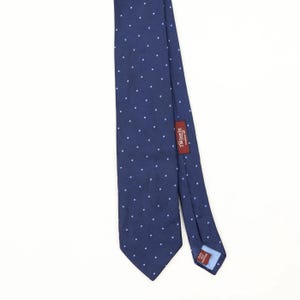 Pode incluir: Uma gravata azul-marinho com um padrão repetitivo de pequenas bolinhas brancas. A gravata inclui uma etiqueta de tecido vermelho costurada na parte de trás com o texto TM Lewin Established 1898 e uma etiqueta menor na ponta com Pure Silk Hand Made.