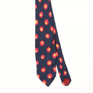 May include: A dark navy blue necktie featuring a repeating pattern of circular motifs. Each motif consists of a bright yellow centre surrounded by a larger red ring. The tie has a pointed end and a smooth fabric texture, displaying a consistent design.