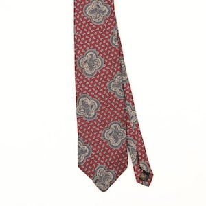 Può includere: Una cravatta rossa caratterizzata da un motivo ripetitivo di piccole forme paisley blu scuro, intervallate da motivi quadrifogli più grandi e chiari che contengono intricati disegni paisley blu e beige su tutta la superficie.