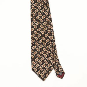Pode incluir: Uma gravata com fundo castanho escuro e um padrão repetitivo de formas paisley em tons de bege, azul e creme. A gravata inclui uma pequena etiqueta preta na parte de trás com texto branco que diz Made in U.S.A. e 100% Silk.