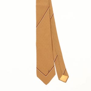 Peut inclure: Une cravate beige ornée d'un motif discret de petits points sombres sur le tissu. La cravate présente un détail de rayure diagonale avec une fine ligne rouge foncé et une bordure pointillée. Un petit monogramme brodé est visible près de la pointe.