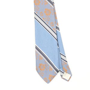 Op de afbeelding: Een das met een lichtblauwe achtergrond, een patroon van oranje bloemen en diagonale strepen in wit en donkerblauw. De das heeft een puntig uiteinde en een zichtbaar wit stoffen label aan de achterkant bij het smalle uiteinde.
