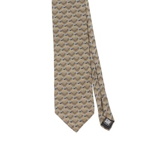 May include: A tan necktie featuring a repeating geometric pattern of small, light blue squares arranged within a series of interlocking chevron shapes. The tie includes a black fabric label on the back with white text detailing care instructions.