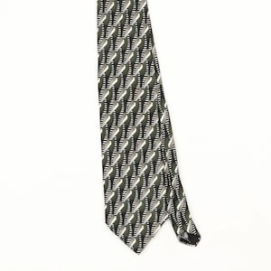 May include: A necktie featuring a repeating geometric pattern of olive green, black, and white diagonal stripes. The design consists of angled segments that create a textured, layered appearance across the entire length of the fabric accessory.