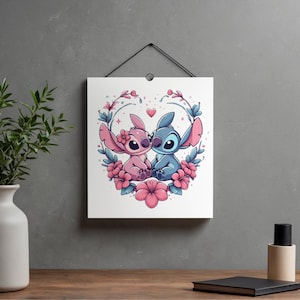 May include: A square wall hanging featuring a cartoon illustration of two alien creatures, one pink and one blue, sitting together. They are surrounded by a circular arrangement of pink hibiscus flowers and blue leaves, with a small pink heart above them.