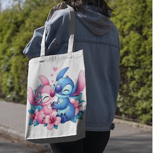May include: A white canvas tote bag featuring a colorful graphic print of two cartoon characters, one blue and one pink, surrounded by pink and blue hibiscus flowers with small pink hearts floating above them, held by two fabric shoulder straps.
