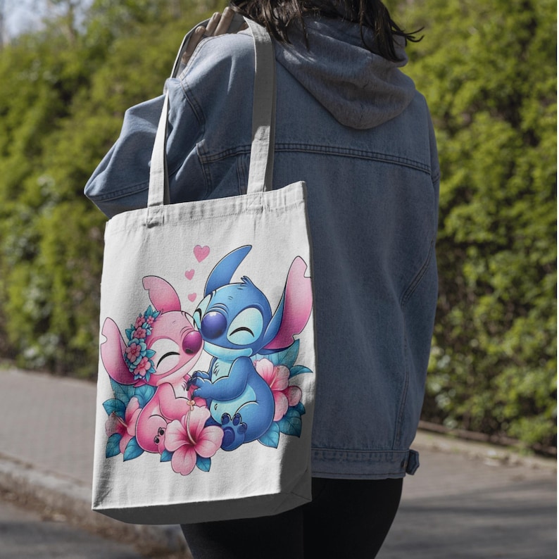 May include: A white canvas tote bag featuring a colorful graphic print of two cartoon characters, one blue and one pink, surrounded by pink and blue hibiscus flowers with small pink hearts floating above them, held by two fabric shoulder straps.