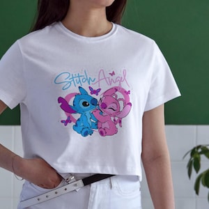 May include: A white short-sleeved t-shirt featuring a graphic print of the blue character Stitch and the pink character Angel. The design includes a pink heart outline, small purple butterflies, and the text Stitch Angel in blue and pink script.