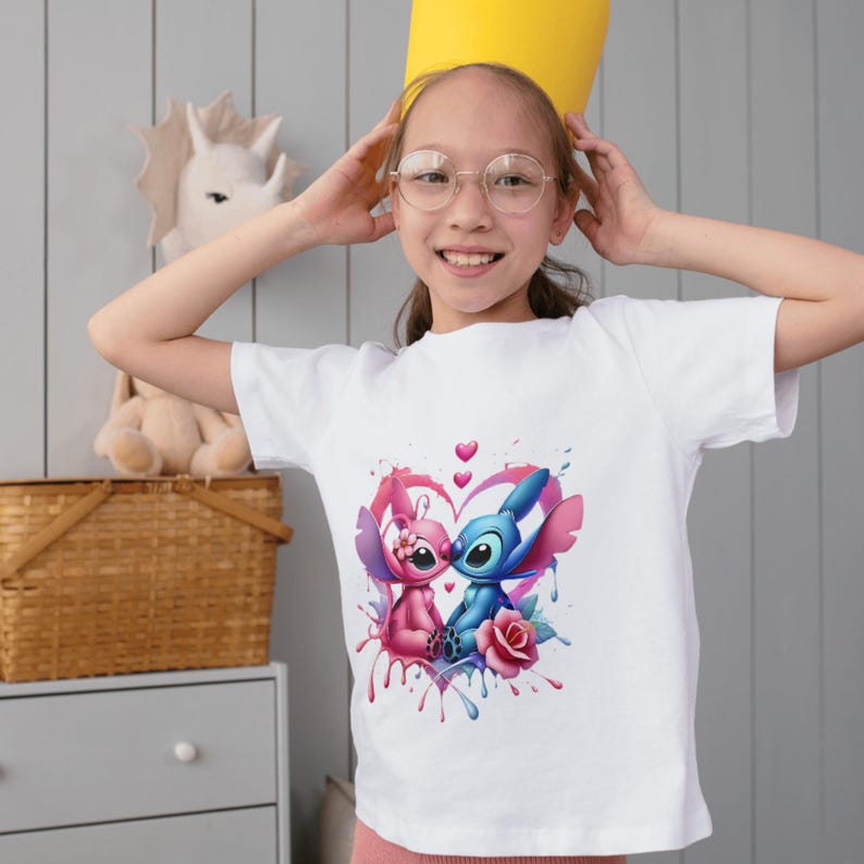 May include: A white short-sleeved t-shirt featuring a graphic print of two blue and pink alien characters sitting together inside a heart shape, surrounded by pink and blue paint drips and a single pink rose at the bottom center of the design.