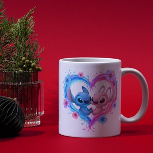 May include: A white ceramic mug featuring a graphic of two cartoon characters, one blue and one pink, positioned inside a heart shape formed by blue and pink liquid splashes and colorful hibiscus flowers against a solid red background.