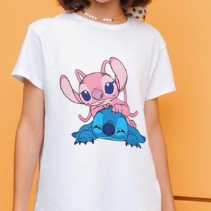 May include: A white short-sleeved t-shirt featuring a graphic print of a pink alien character sitting on top of a blue alien character. The design is centered on the chest area of the shirt, showcasing the two characters with large ears and eyes.