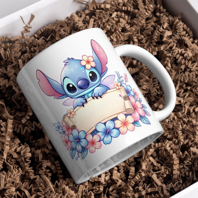 May include: A white ceramic mug featuring a blue alien character with large ears and a flower on its head, holding a blank beige scroll. The design is surrounded by a cluster of pink and blue tropical flowers and green leaves on a white background.