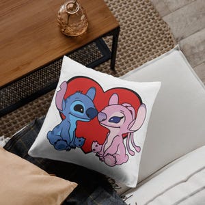 May include: A square white throw pillow featuring a graphic illustration of two cartoon characters, one blue and one pink, sitting in front of a large red heart. The characters have large ears and dark eyes, depicted in a simple, clean line art style.