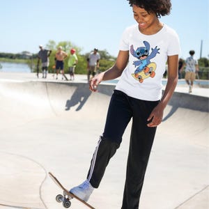 May include: A white short-sleeved t-shirt featuring a graphic print of the blue character Stitch from Lilo and Stitch riding a yellow skateboard with orange wheels. The shirt is paired with black athletic pants with white side stripes.