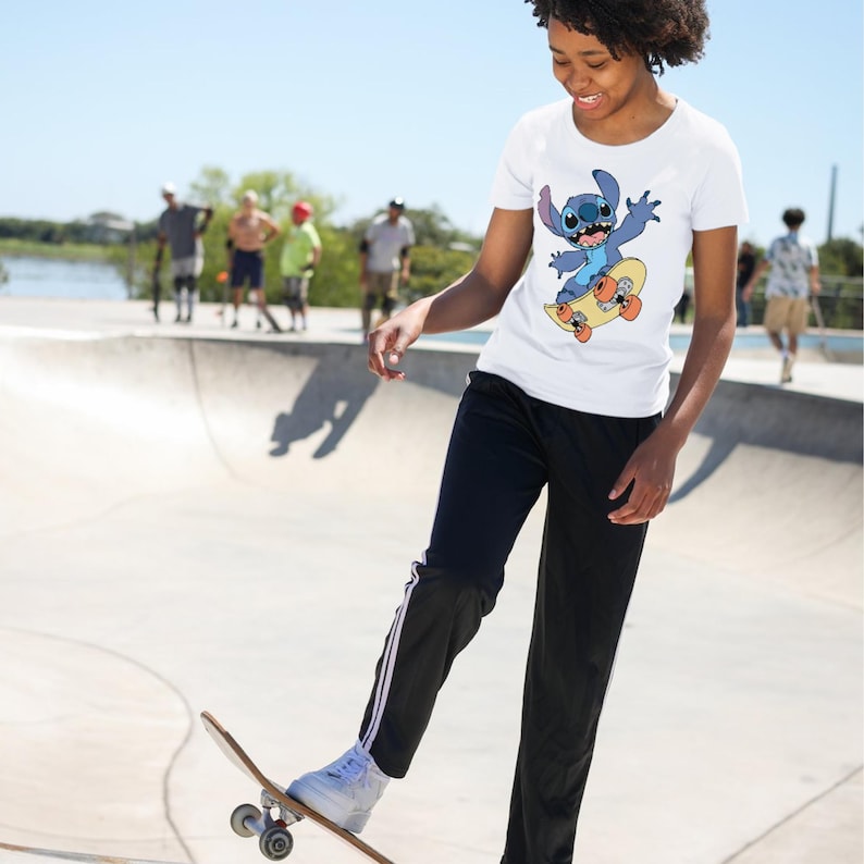 May include: A white short-sleeved t-shirt featuring a graphic print of the blue character Stitch from Lilo and Stitch riding a yellow skateboard with orange wheels. The shirt is paired with black athletic pants with white side stripes.