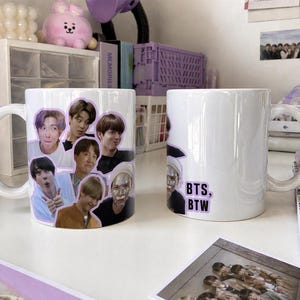 Può includere: Due tazze di ceramica bianca. Una tazza presenta un collage di volti, l'altra ha il testo "BTS, BTW" in un rettangolo viola. Entrambe le tazze hanno un manico bianco e sono su una superficie bianca.