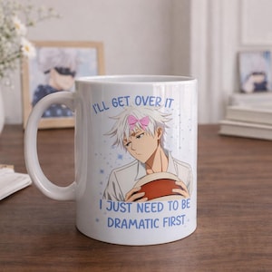 Può includere: Tazza in ceramica bianca con un'illustrazione di un personaggio anime e la scritta "I'll get over it. I just need to be dramatic first." La tazza ha un manico curvo ed è appoggiata su una superficie di legno. Le margherite in un vaso bianco sono sullo sfondo.