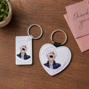 Peut inclure: Deux porte-clés avec des illustrations de personnages d'anime. L'un est rectangulaire, l'autre en forme de cœur. Les deux présentent un personnage aux cheveux blancs et rouges, une veste bleue et le texte "tryna act like I'm not crazy as hell."