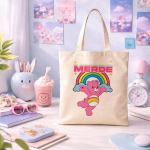 Può includere: Borsa tote beige con un personaggio di Care Bear rosa con un arcobaleno e la parola "MERDE" in rosa e verde. La borsa ha due manici ed è appoggiata su una scrivania bianca. Lo sfondo include elementi decorativi.