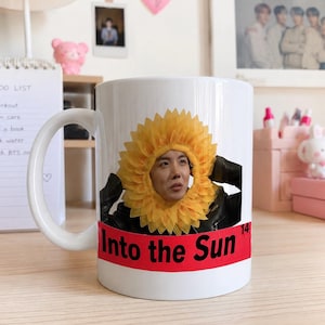 May include: A white ceramic mug featuring a graphic of a person wearing a yellow sunflower headpiece. Below the image, a red horizontal band contains the black text "Into the Sun" with the small number 14 positioned at the far right edge.