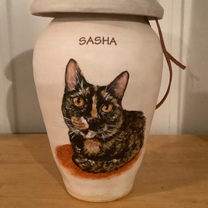 May include: White ceramic pet urn with a brown leather strap. The urn has a painted portrait of a tabby cat with green eyes and the name "SASHA" written on the front.