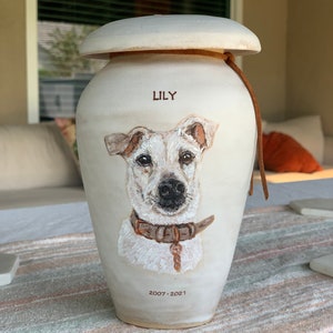 May include: White ceramic pet urn with a painted portrait of a Jack Russell Terrier. The urn has a lid and a brown leather cord. The dog's name "LILY" and the dates "2007-2021" are written on the urn.