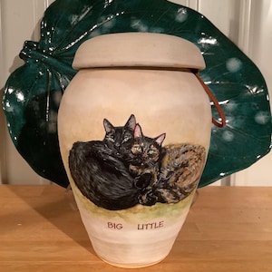 May include: Ceramic pet urn with a hand-painted image of two black cats cuddling. The urn is cream-colored with a brown lid and a leather strap. The words "BIG" and "LITTLE" are written on the front of the urn.