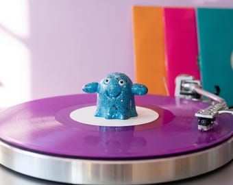 May include: A blue speckled figurine with a rounded head, two side arms, and a smiling face sits on the center of a vibrant purple vinyl record. The record is placed on a silver turntable with a metallic tone arm and a white center label.
