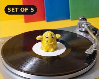 May include: A yellow ceramic ghost-shaped record stabilizer with white and blue paint splatters, two googly eyes, and a smiling mouth. The object sits on a black vinyl record on a silver turntable. A black label reads SET OF 5 in yellow text.