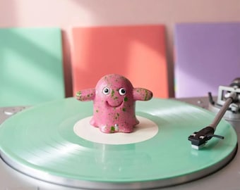 May include: A pink record stabilizer with green speckles, featuring a small creature-like shape with two arms, a smiling mouth, and two white eyes with black pupils, positioned in the center of a light green vinyl record on a turntable.