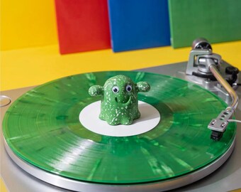 May include: A small green ghost-shaped figure with white speckles and googly eyes sits on the center of a translucent green marbled vinyl record. The record is placed on a silver turntable platter with a metal tonearm positioned to the right side.