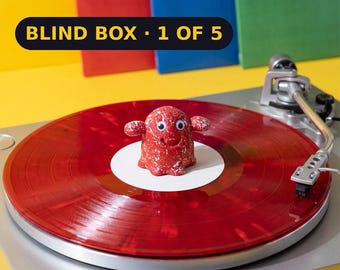 May include: A red, speckled, ghost-shaped figurine with googly eyes sits on a translucent red vinyl record on a silver turntable. A black banner with yellow text reads BLIND BOX - 1 OF 5. The background features yellow, red, blue, and green panels.