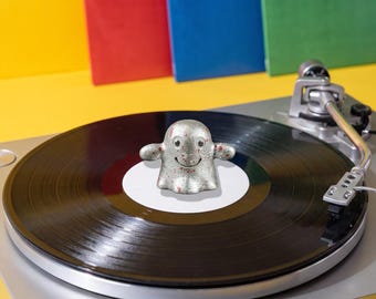 May include: A silver-colored ghost figurine with a speckled finish and small red accents sits on the center of a black vinyl record. The record is placed on a silver turntable with a tone arm visible on the right side of the device.