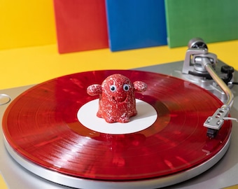 May include: A small red speckled figurine with googly eyes sits on a translucent red vinyl record placed on a silver turntable. The record features a white center label. The figurine has two small arms extended to the sides and a smiling mouth.