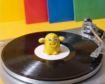 May include: A small yellow ceramic figurine with a speckled pattern and googly eyes, shaped like a creature with outstretched arms, positioned on the center of a black vinyl record resting on a silver turntable with a metallic tone arm.