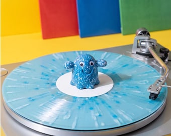 May include: A light blue vinyl record with a white splatter pattern sits on a silver turntable. A small, blue, speckled, ghost-shaped figure with two googly eyes and a smiling mouth is positioned in the center of the record on the white label.