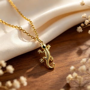 May include: A gold-toned necklace featuring a small, detailed lizard pendant. The pendant is shaped like a gecko with textured scales and a curled tail, suspended from a delicate gold-toned chain link. The metal has a polished, reflective finish.